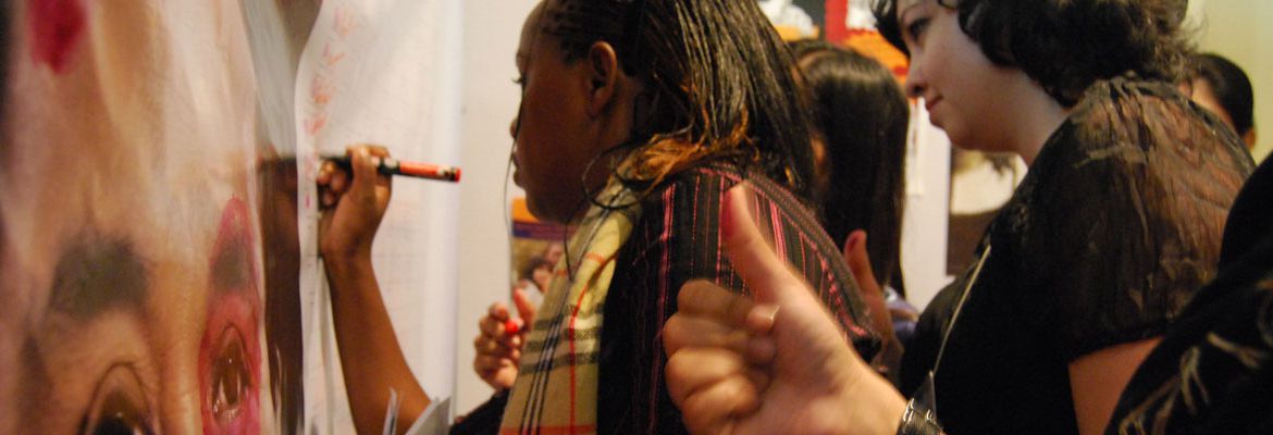 A black woman writing something on a sheet of paper hung on a wall. Around her are more people with markers, or thumbs with red ink on them waiting for their turn.