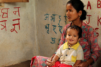 Woman wearing a pink and blue Indian suit sitting cross-legged on floor, with a toddler on her lap. The baby is wearing a yellow shirt and red shorts. Behind them on the wall are both the English and Hindi alphabets.
