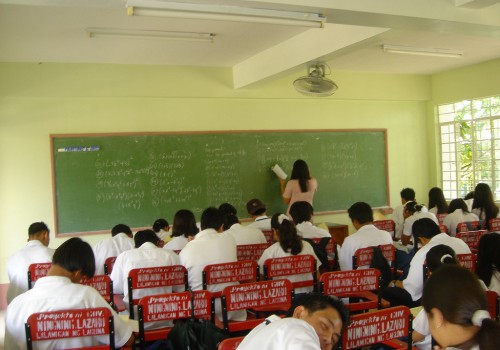 Classroom. A teacher writing on a board, and girls and boys in white school uniform are sitting looking down at their books.