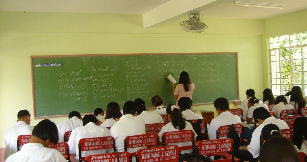 Classroom. A teacher writing on a board, and girls and boys in white school uniform are sitting looking down at their books.