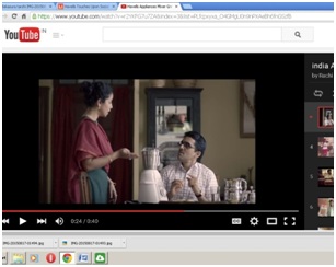 Screenshot of a YouTube page. The video still shows a husband sitting at a dining table. His wife stands nearby, talking to him, pointing to a mixer kept on the table.