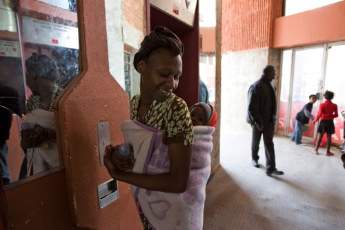 An African woman carrying a baby on her back with the help of a cloth tied to her. She is carrying a milk bottle, and is trying to look behind her shoulder at the baby.