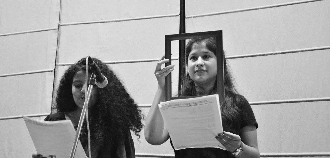 Black and white photo of two young women speaking on a stage, reading out from the sheets of paper in their hands. One wears a saree, and has curly hair left open. The other wears a top, and has her hair open falling till her chest. She holds a rectangle-shaped structure in front of her face through which she sees across.
