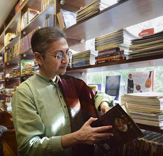 Lata Mani in a bookshop. She has short hair and is wearing a green kurta and glasses. She flips through a book by a shelf.