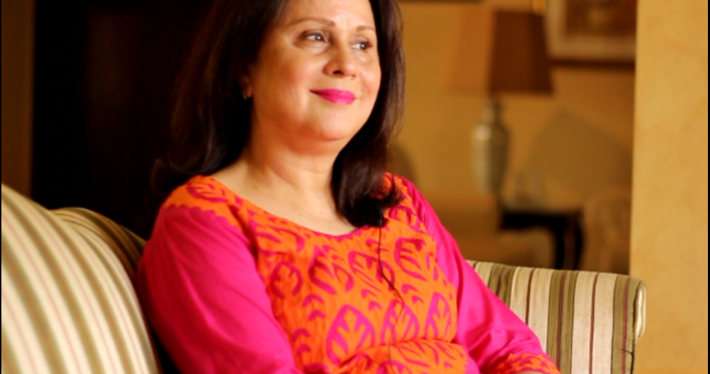 Uzma Noorani, a human rights activist, sitting on a sofa smiling. She is wearing an orange and pink Indian suit, and has her hair falling on her shoulders.