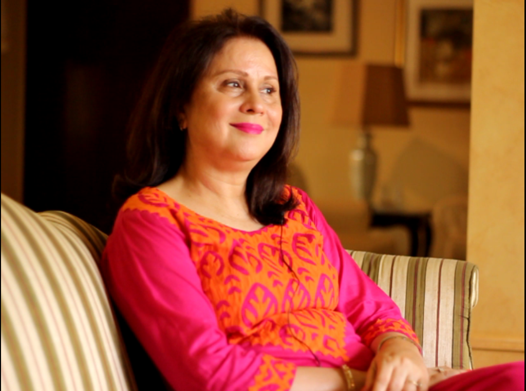 Uzma Noorani, a human rights activist, sitting on a sofa smiling. She is wearing an orange and pink Indian suit, and has her hair falling on her shoulders.