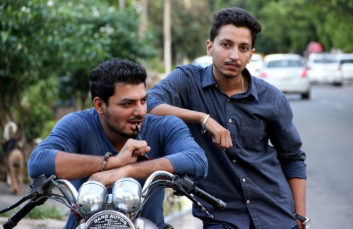 Two men hanging out on a side of a road, by their motorcycle. One wearing a blue tee sits on the motorcycle with his elbows on the handle. The other, in a blue shirt, stands resting his elbows on the first one's shoulder.