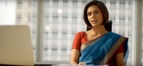 A young woman in a blue saree and red blouse sitting in her office chair, with a laptop open in front of her. Skyscrapers are visible through the glass walls behind her. She has a short haircut.