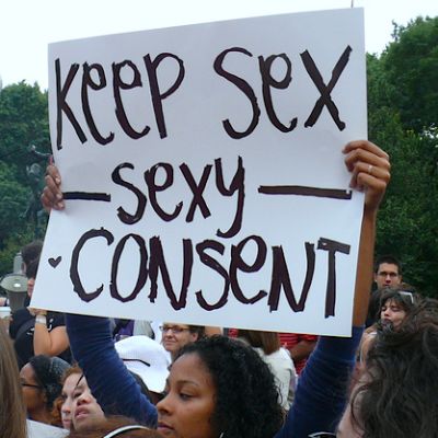 A brown woman in a blue top standing amongst a crowd raises a placard that reads in black on a white background, "Keep sex sexy - consent."