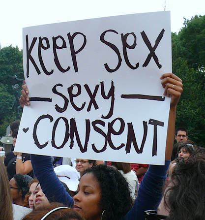 A brown woman in a blue top standing amongst a crowd raises a placard that reads in black on a white background, "Keep sex sexy - consent."