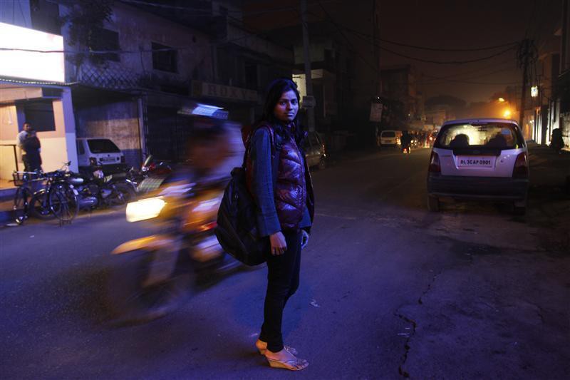 An Indian woman on the middle of a road at night. A motorcycle whooshes past her. She is wearing a blue jacket, black jeans, sandals, and is carrying a handbag on her shoulder. Her hair fall on her shoulders.
