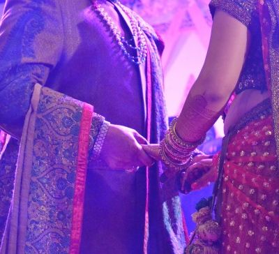 A bride and her mother holding hands face-to-face during the wedding celebration. Their faces are cut from the picture - we can see from their neck to their knees, dressed in a bride's lehnga, and a heavy Indian suit.