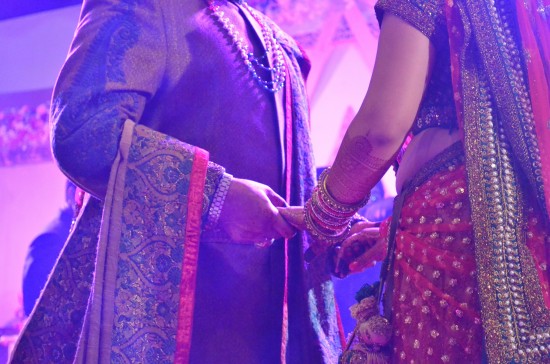 A bride and her mother holding hands face-to-face during the wedding celebration. Their faces are cut from the picture - we can see from their neck to their knees, dressed in a bride's lehnga, and a heavy Indian suit.