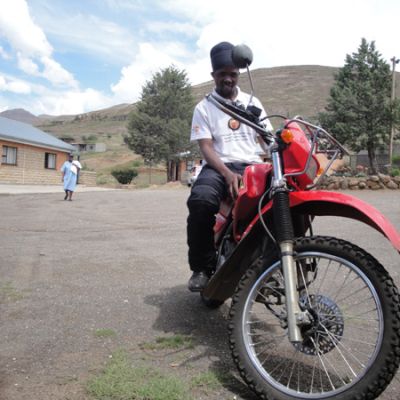 A black man in a white shirt and black pants sitting on a motorcycle in a peaceful hilly town.