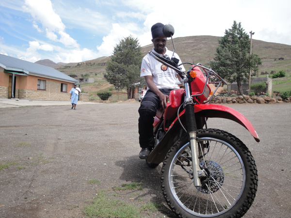 A black man in a white shirt and black pants sitting on a motorcycle in a peaceful hilly town.