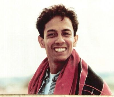 Dominic D’Souza, a young gay man. He is smiling at the camera. He wears a white vest peeking through this black-and-white striped shirt, and a red shawl thrown around his shoulders.
