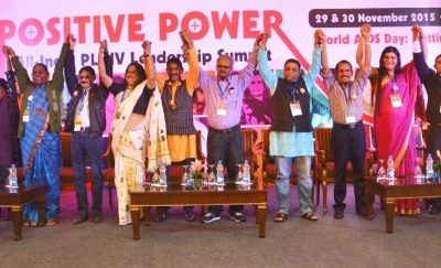 Professionals standing in a row on stage. They join their hands and raised them up in celebration. The poster on the wall behind them reads, "Positive power".