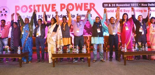 Professionals standing in a row on stage. They join their hands and raised them up in celebration. The poster on the wall behind them reads, "Positive power".