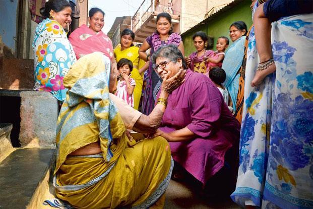 Meena Seshu squatting on the floor as an older woman sitting on the floor caresses her on the cheek. Several women stand around them, looking on. Seshu is wearing a purple kurta, and the older woman wears a yellow saree.