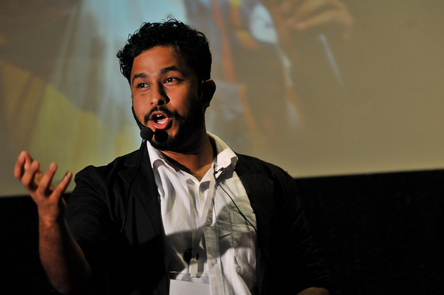 Abish Matthew in a white shirt and black coat speaking into ear mike on a stage.