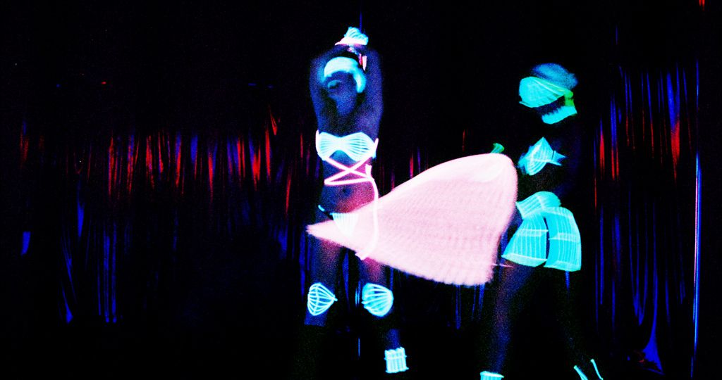 Two women dancing in a strip club. Their clothes shine in green neon light in the dark background.