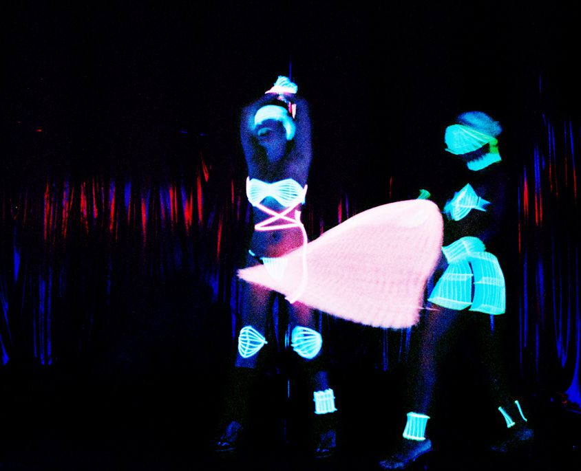 Two women dancing in a strip club. Their clothes shine in green neon light in the dark background.
