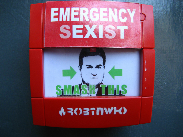 In the vein of an emergency fire alarm, "emergency sexist" is written in white on a red instrument. "Smash this" is written in green, pointing to the face of a man behind a glass.