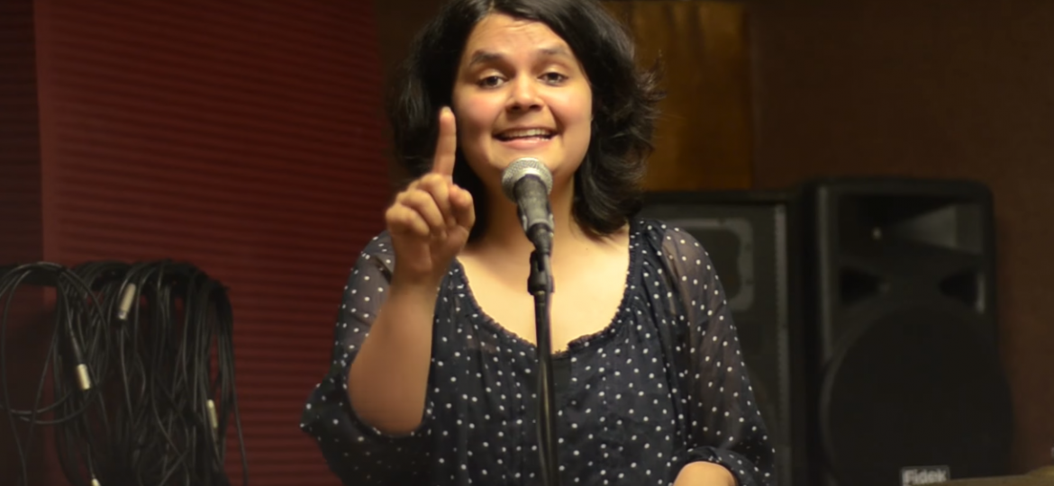 Picture of Priyam Redican standing in front of a mic. She is wearing a black top and has short shoulder-length hair