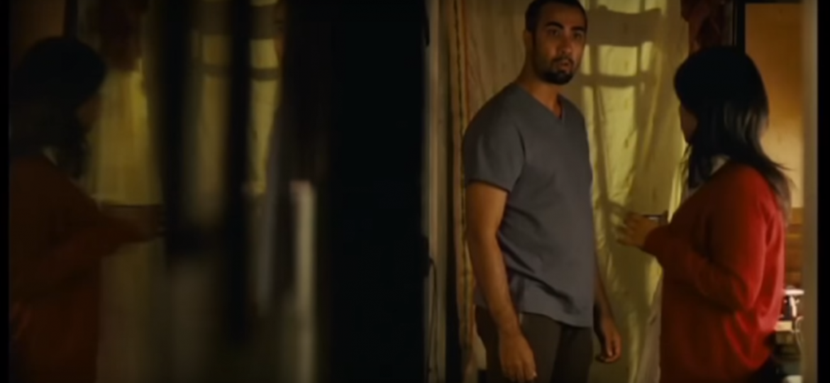 A man in a grey tee shirt and black pajamas standing, looking surprised at something.