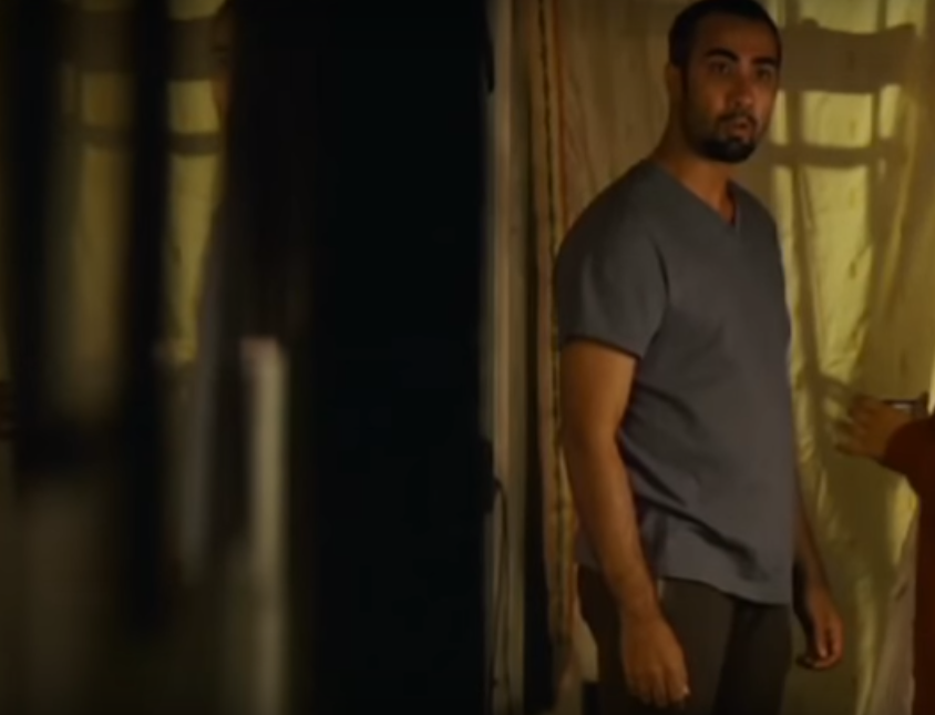 A man in a grey tee shirt and black pajamas standing, looking surprised at something.