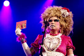 Picture of a Japanese drag queen. She wears a big blonde wig and holds up a pink card.