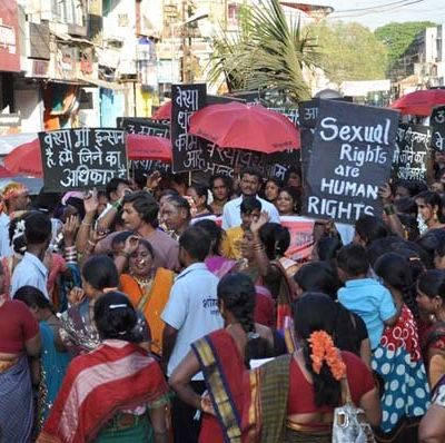 A protest by sex workers. They are wearing sarees, holding red umbrellas, and placards reading "Stop policing our morals", "Sexual rights are human rights".