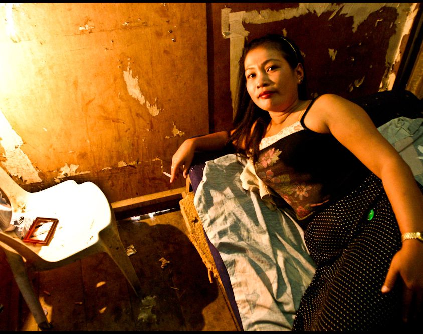 A sex worker lying on her side on a bed in a run-down room, smoking.