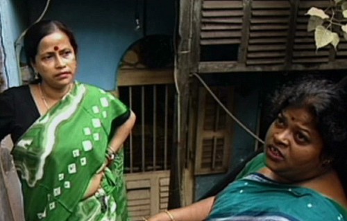 Two sex workers standing and talking outside a chawl. They both are dressed in sarees, and are wearing a red bindi on their forehead.