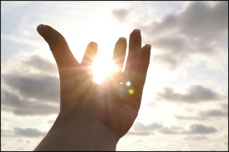 Photo of a hand in front of a cloudy and sunny sky. The picture is clicked such that it seems the sun is held in the palm of the hand.