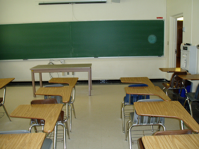 Photo of an empty classroom. Chairs, desks, teacher's table and a clean blackboard are visible.