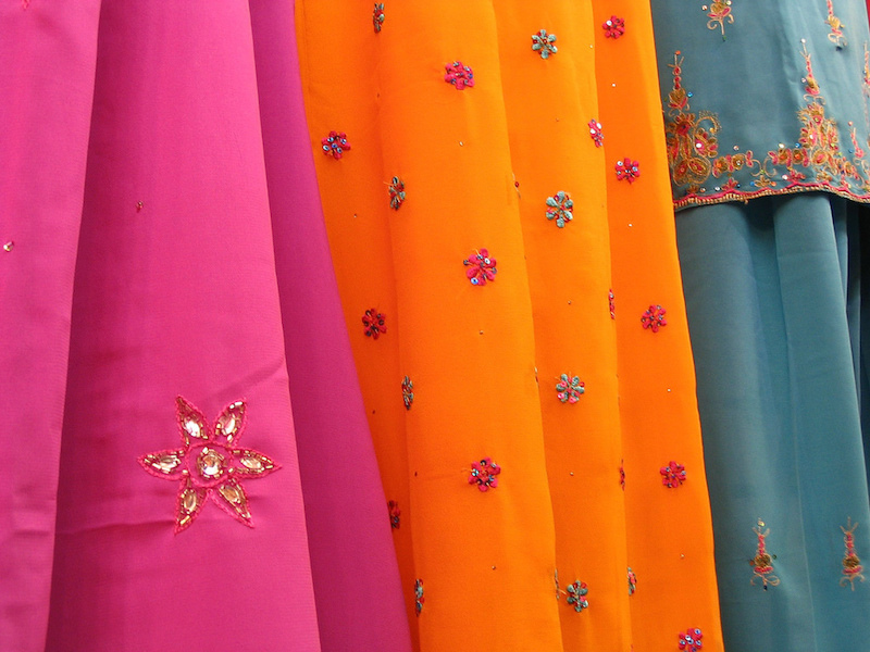 Photo of Indian dresses. A pink saree, an orange saree, and a blue salvar suit - all three have colourful flowers and designs on them. Screen reader support enabled. Photo of Indian dresses. A pink saree, an orange saree, and a blue salvar suit - all three have colourful flowers and designs on them.
