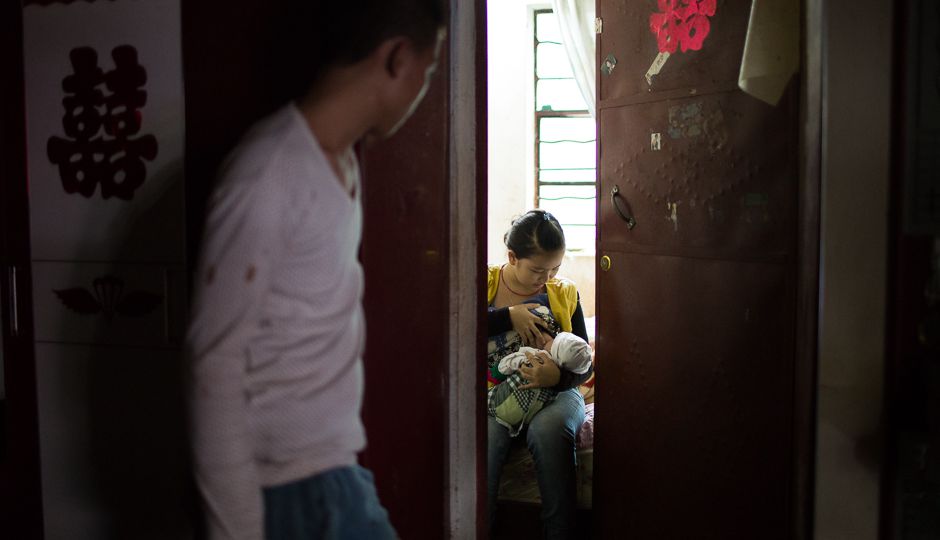 A boy looking in a room through a creaked door. There, a girl is breastfeeding a baby.