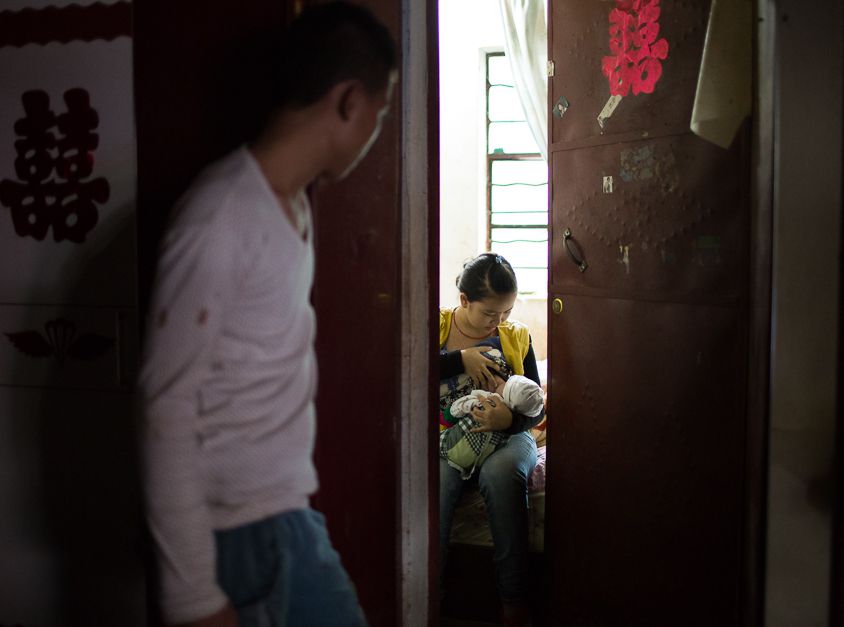 A boy looking in a room through a creaked door. There, a girl is breastfeeding a baby.