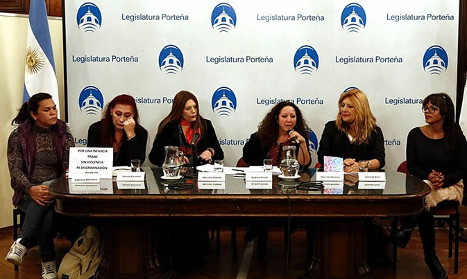 Six women behind a table giving a press conference. To the left stands the Aragentinian flag. Behind them is a white cloth on which is printed "legislatura portena" with a symbol or logo of Buenos Aires City Legislature in blue colour. The words and the photo are printed all over the cloth in a line.