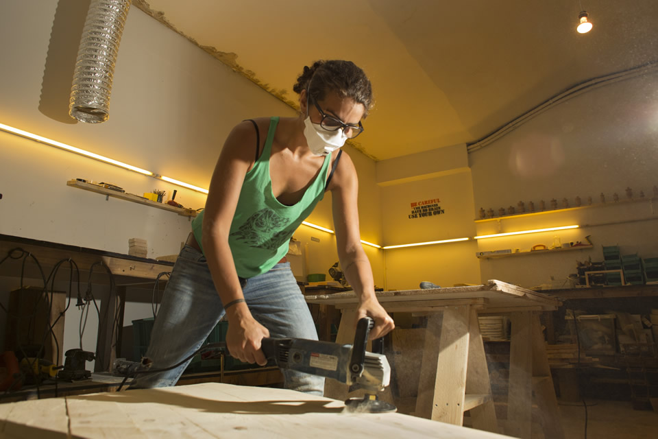 A woman in a green tank-top and light blue jeans doing carpentary. Hher hair are tied up, she is wearing black-framed glasses, and a white mask on the mouth.