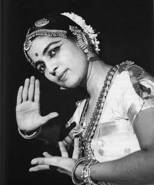 Black-and-white photo of Indian bharatnatyma dancer Rukmini Devi holding a mudra. She is wearing traditional dance clothing with flowers in her head, and jewellery on the forehead, ears, and neck. The image is from the chest up.