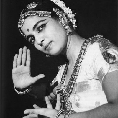 Black-and-white photo of Indian bharatnatyma dancer Rukmini Devi holding a mudra. She is wearing traditional dance clothing with flowers in her head, and jewellery on the forehead, ears, and neck. The image is from the chest up.