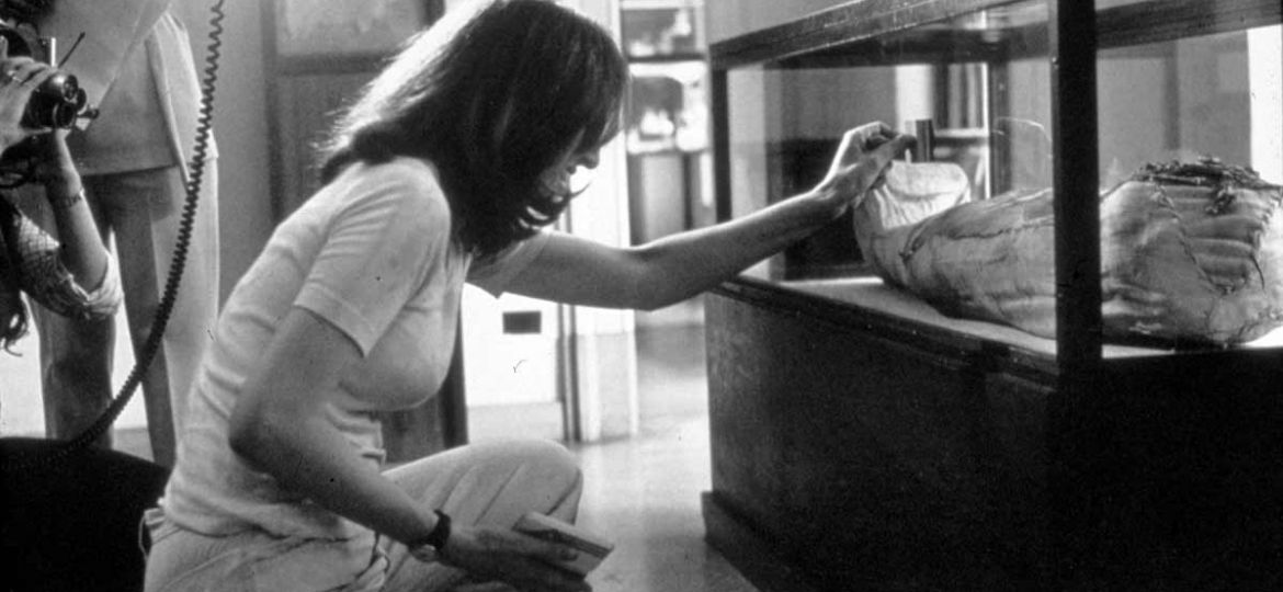 A black-and-white photo of a woman in jeans and tee-shirt bent down to clean a glass vitrine in a museum.