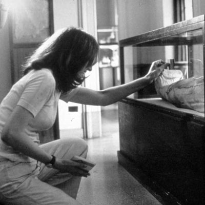 A black-and-white photo of a woman in jeans and tee-shirt bent down to clean a glass vitrine in a museum.