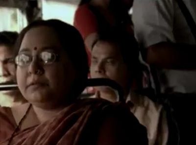 Screenshot from a video. A middle-aged Indian woman sitting in a crowded bus. She is wearing orange saree, red bindi, and specks.