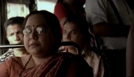 Screenshot from a video. A middle-aged Indian woman sitting in a crowded bus. She is wearing orange saree, red bindi, and specks.