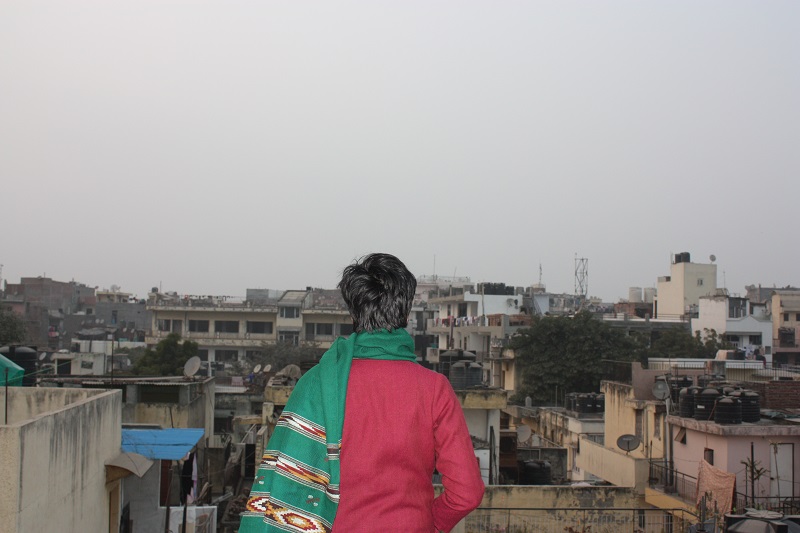Back of a woman with boy-cut hair, wearing red sweater, and a green shawl round her neck, looking at the view from a terrace. She can see houses and more houses.