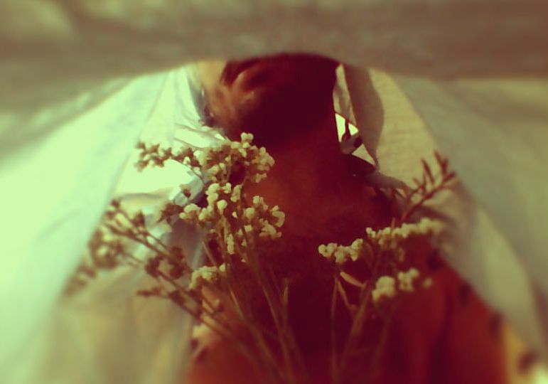 A nude man with a bedsheet over his head, is holding a bunch of small white flowers. The photo is clicked from inside the bedsheet. He is visible from the lips till the midrif.