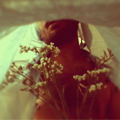 A nude man with a bedsheet over his head, is holding a bunch of small white flowers. The photo is clicked from inside the bedsheet. He is visible from the lips till the midrif.